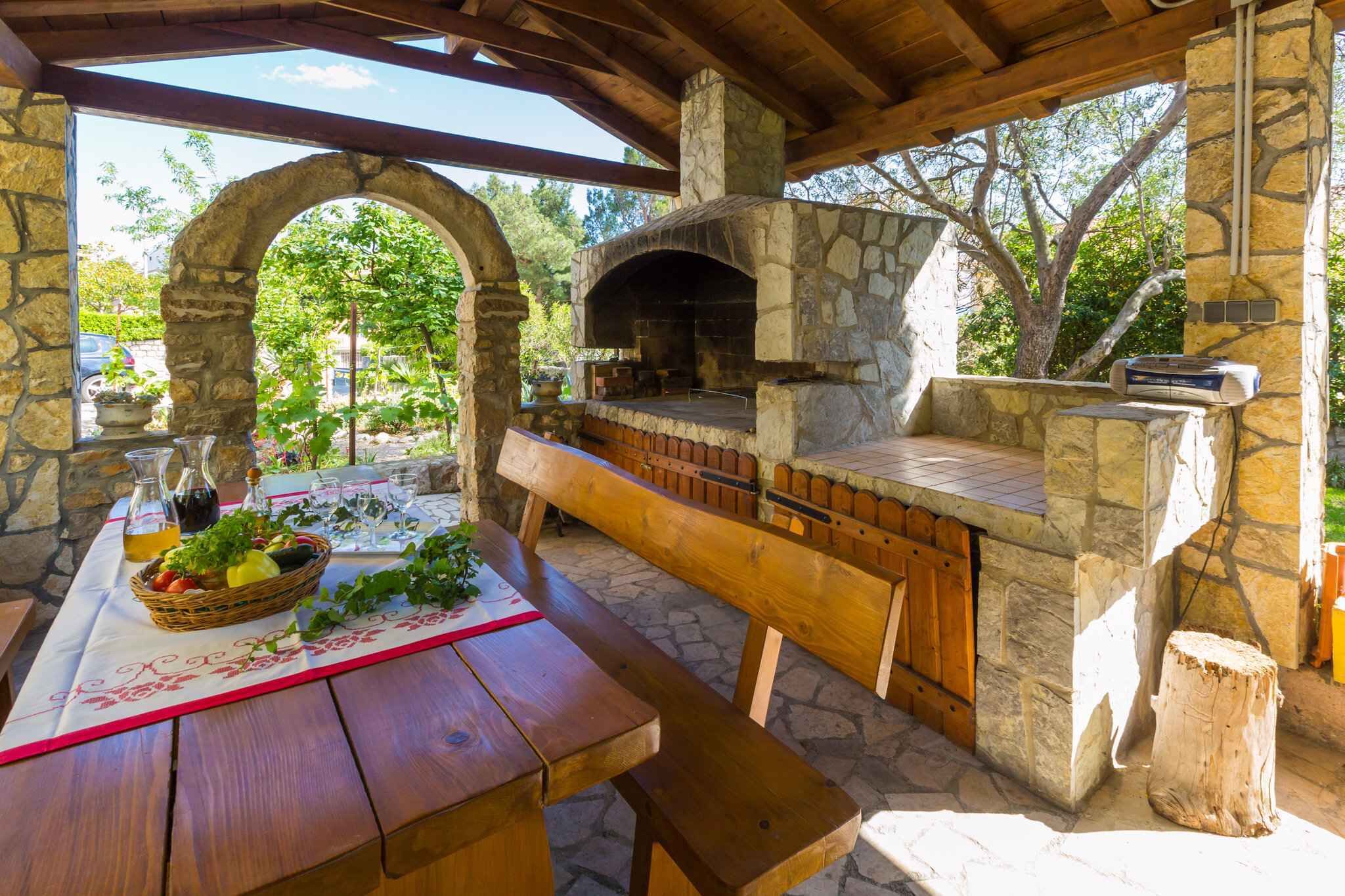 An outdoor stone oven with a wooden table and benches, surrounded by stone walls and a covered area.