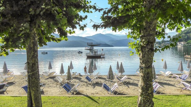 Una playa junto al lago con sillas de descanso y sombrillas, una torre de salvavidas y montañas al fondo.