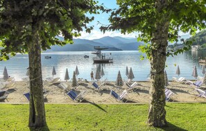 A lakeside beach with lounge chairs and umbrellas, a lifeguard tower, and mountains in the background.
