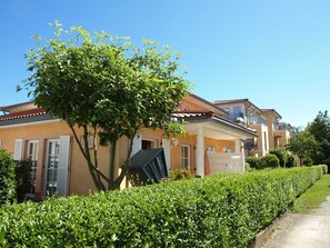 Ein Wohngebäude mit Balkon, einem Baum im Vordergrund und einer gepflegten Hecke.