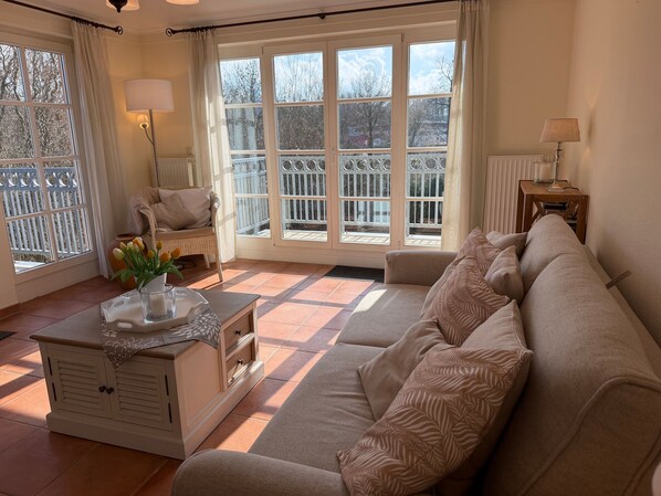 Una sala de estar con un sofá, una mesita de noche con una lámpara, una silla, un ventanal con vistas a un balcón y una pequeña mesa con un florero y una vela.
