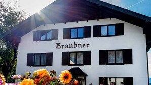 Un bâtiment blanc dont la façade porte l’inscription « Brandner », avec de nombreuses fenêtres et un jardin fleuri au premier plan.