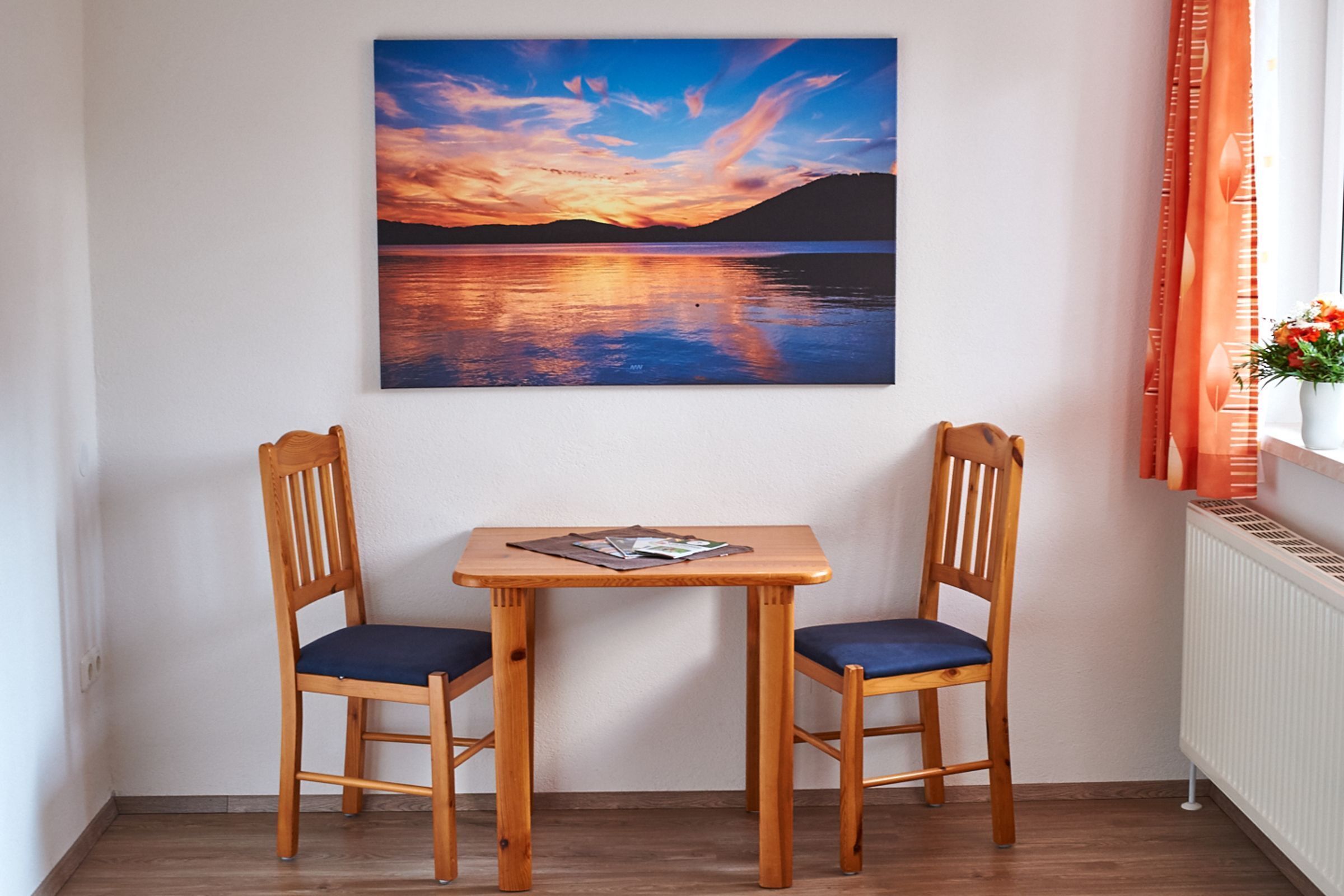 Una habitación con una mesa de madera y dos sillas, un cuadro de un atardecer sobre el agua, y una ventana con cortinas.