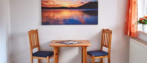 Una habitación con una mesa de madera y dos sillas, un cuadro de un atardecer sobre el agua, y una ventana con cortinas.