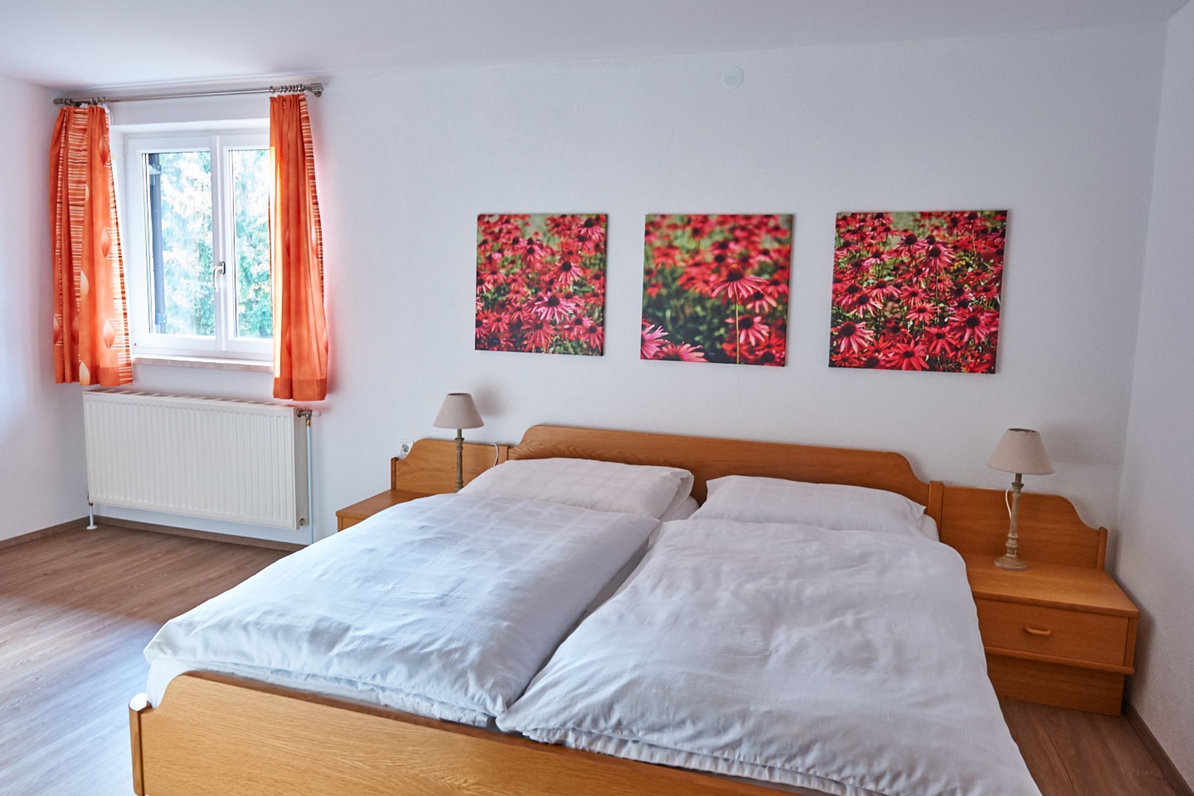 Ein Schlafzimmer mit einem hölzernen Bett, weißer Bettwäsche, einem Fenster mit orangefarbenen Vorhängen und drei floralen Wandbildern.