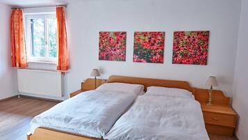 Ein Schlafzimmer mit einem hölzernen Bett, weißer Bettwäsche, einem Fenster mit orangefarbenen Vorhängen und drei floralen Wandbildern.