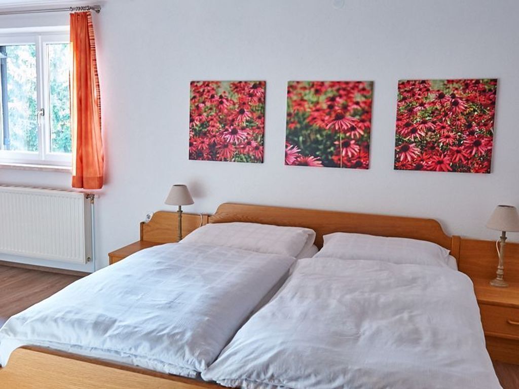 Une chambre à coucher avec un lit en bois, une literie blanche, deux lampes de chevet et trois tableaux représentant des fleurs, accrochés au mur.