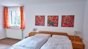 Une chambre avec un lit en bois, du linge de lit blanc, une fenêtre avec des rideaux orange et trois œuvres d’art florales accrochées au mur.
