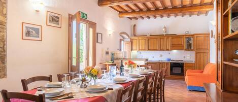 Un comedor con una mesa larga dispuesta para una comida, sillas de madera y una cocina al fondo.