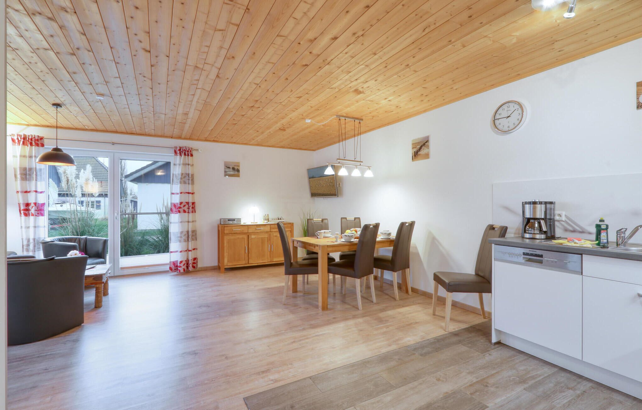 A spacious room with a wooden ceiling, a dining area with a wooden table and chairs, a kitchen with white cabinets, and a large window with curtains.