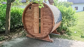 Een houten, vatvormige constructie met een deur, omringd door groen en een geplaveid pad.
