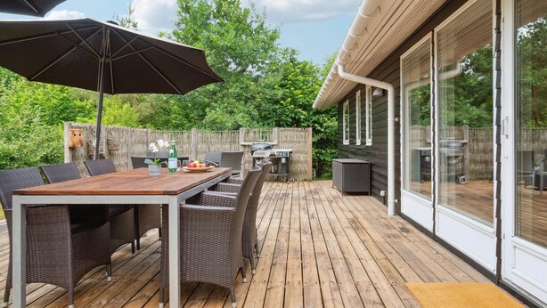 Eine Terrasse im Freien mit Holzdeck, Korbmöbeln, einem Grill und einem großen Sonnenschirm.
