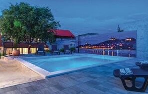 A swimming pool with lounge chairs, a large potted plant, and a view of a cityscape at dusk.