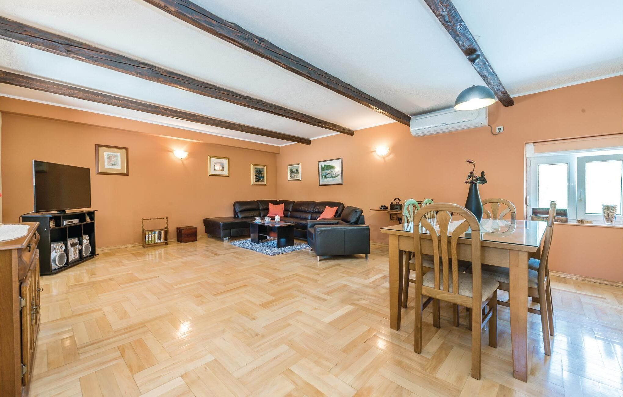 A spacious living room with a wooden herringbone floor, a leather sofa, a dining table with chairs, and a television on a stand.