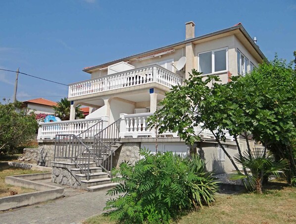 A two-story house with a balcony, stone steps, and surrounding greenery.