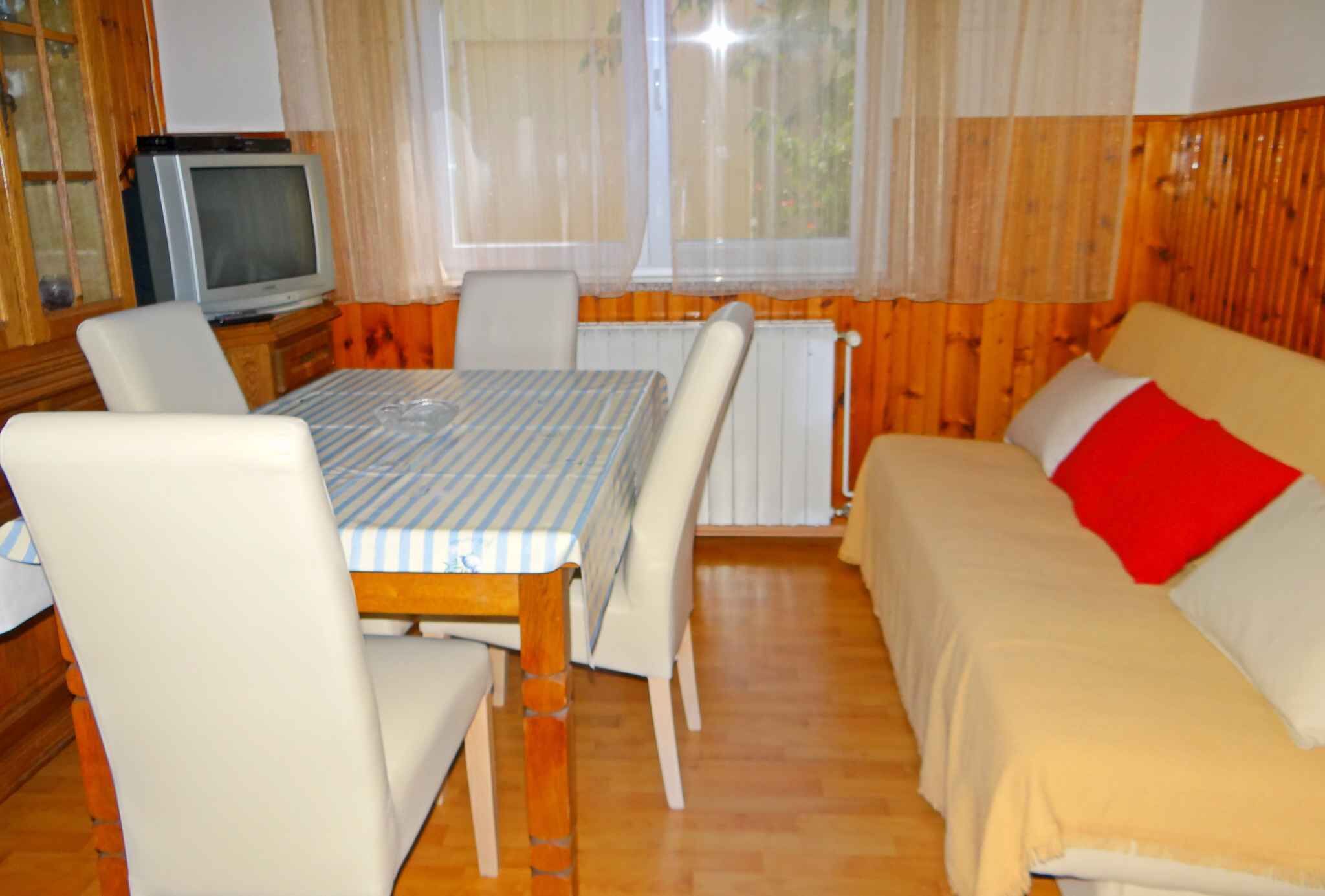 Una habitación con un muro revestido de paneles de madera, un sofá color crema con un cojín rojo, una mesa blanca con mantel a rayas y un televisor sobre un mueble de madera.