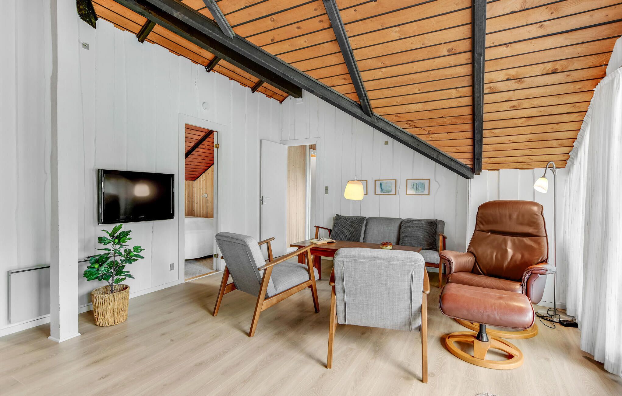 Un salon moderne avec un plafond haut, des poutres en bois, un téléviseur à écran plat, une table à manger avec des chaises, un fauteuil en cuir et une plante en pot.