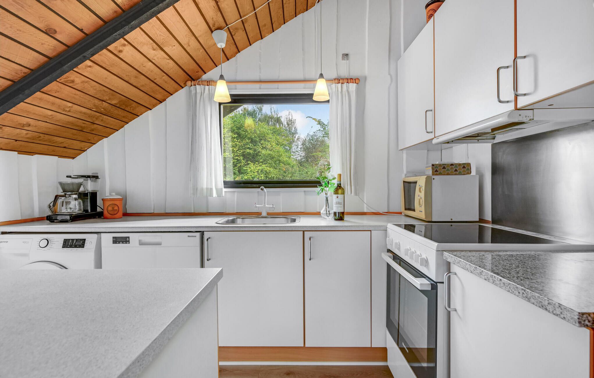 A modern kitchen with white cabinetry, a wooden ceiling, and a window overlooking greenery.