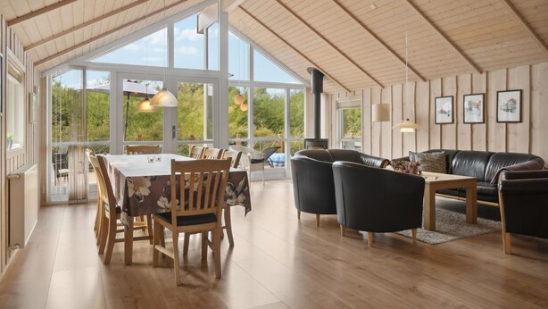 Un vaste espace de vie doté d’un plafond en bois, de grandes fenêtres, d’une cheminée et d’un coin repas avec des meubles en bois.