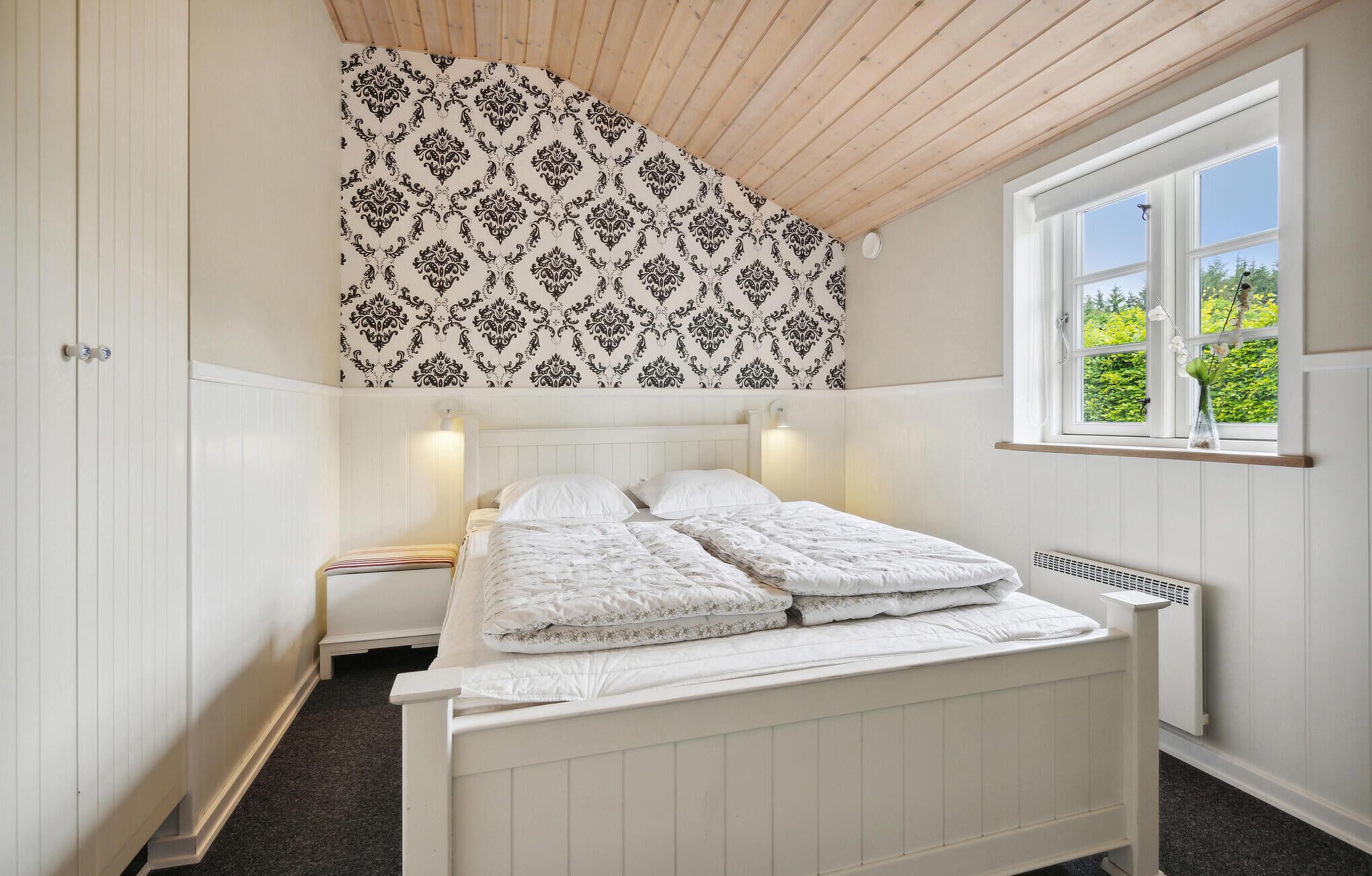 Ein Schlafzimmer mit einem Bett, einem Fenster mit Blick auf Grünflächen und einer Wand mit Muster-Tapete.