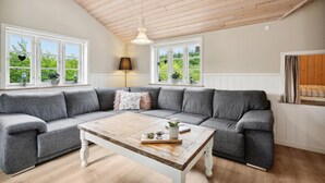 A cozy living room with a grey sectional sofa, a wooden coffee table, and large windows with views of greenery.