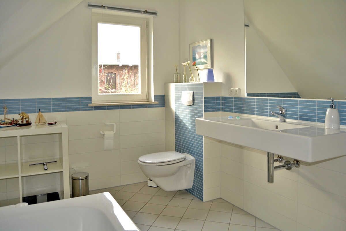 Ein Badezimmer mit Toilette, Waschbecken und Badewanne. Ein Fenster sorgt für natürliches Licht und ein Regal beherbergt dekorative Gegenstände.