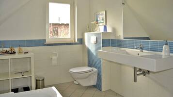 Ein Badezimmer mit Toilette, Waschbecken und Badewanne. Ein Fenster sorgt für natürliches Licht und ein Regal beherbergt dekorative Gegenstände.
