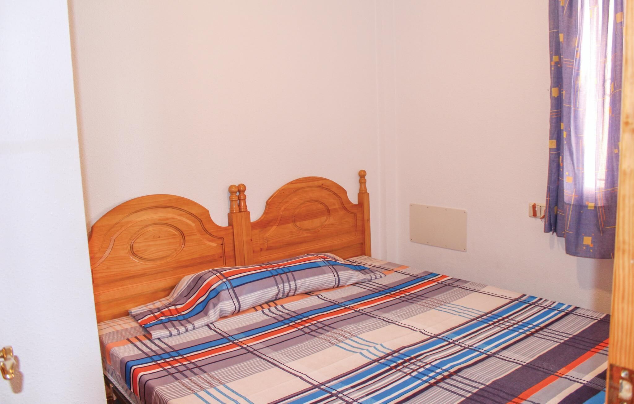 Een enkel bed met een houten hoofdbord, een geruite sprei en een effen muur met een klein lichtschakelaartje en een gordijn dat gedeeltelijk zichtbaar is.