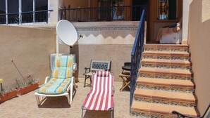 A sunlit outdoor seating area with two lounge chairs, a small table, and a staircase leading to an upper level.