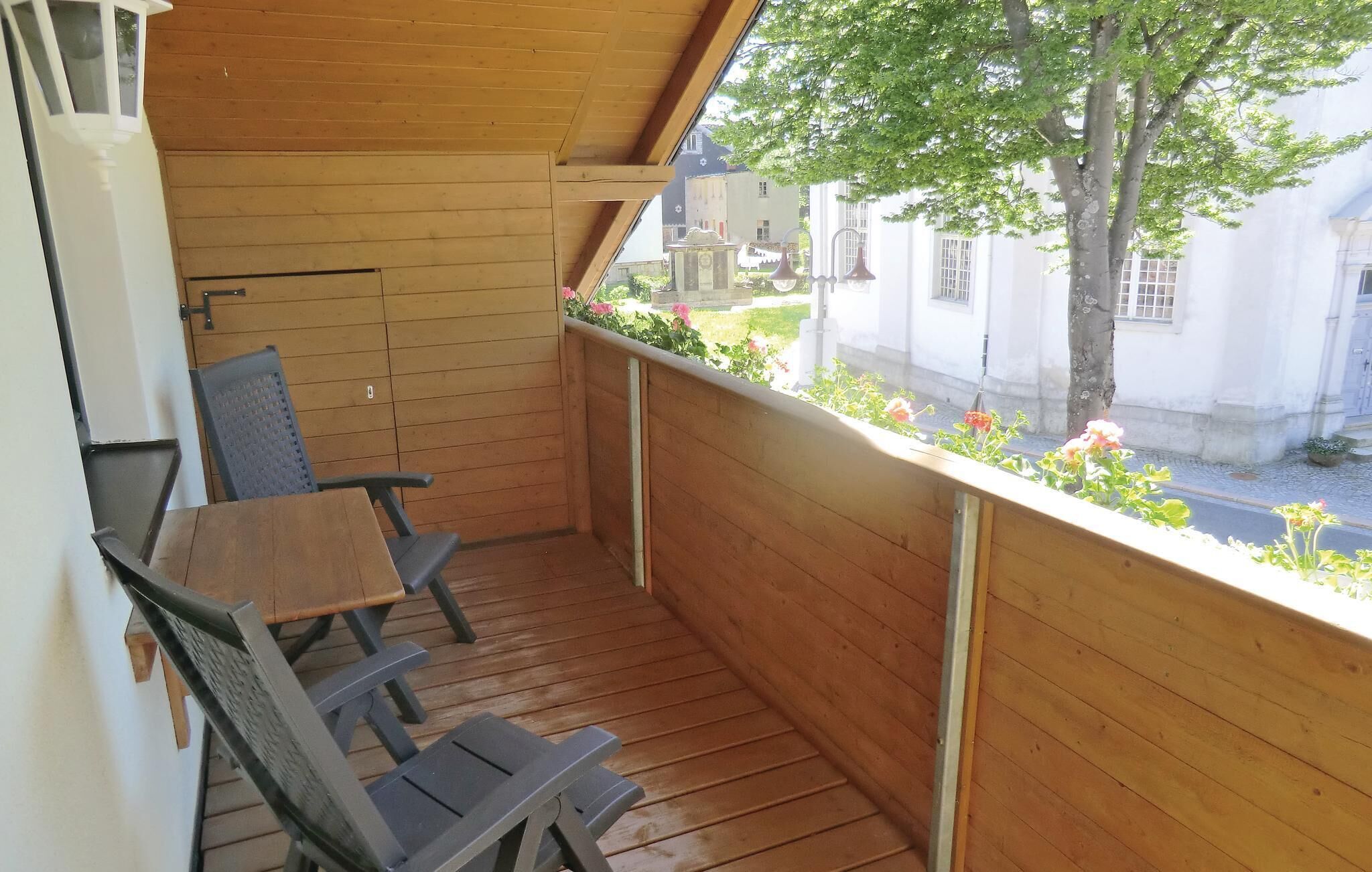 Un balcon en bois avec une table et des chaises, donnant sur une rue bordée d’immeubles et d’un arbre.