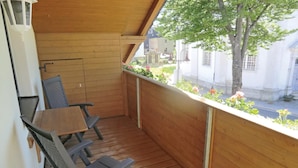A wooden balcony with a table and chairs, overlooking a street with buildings and a tree.
