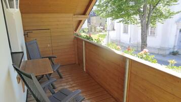 Un balcon en bois avec une table et des chaises, donnant sur une rue bordée d’immeubles et d’un arbre.