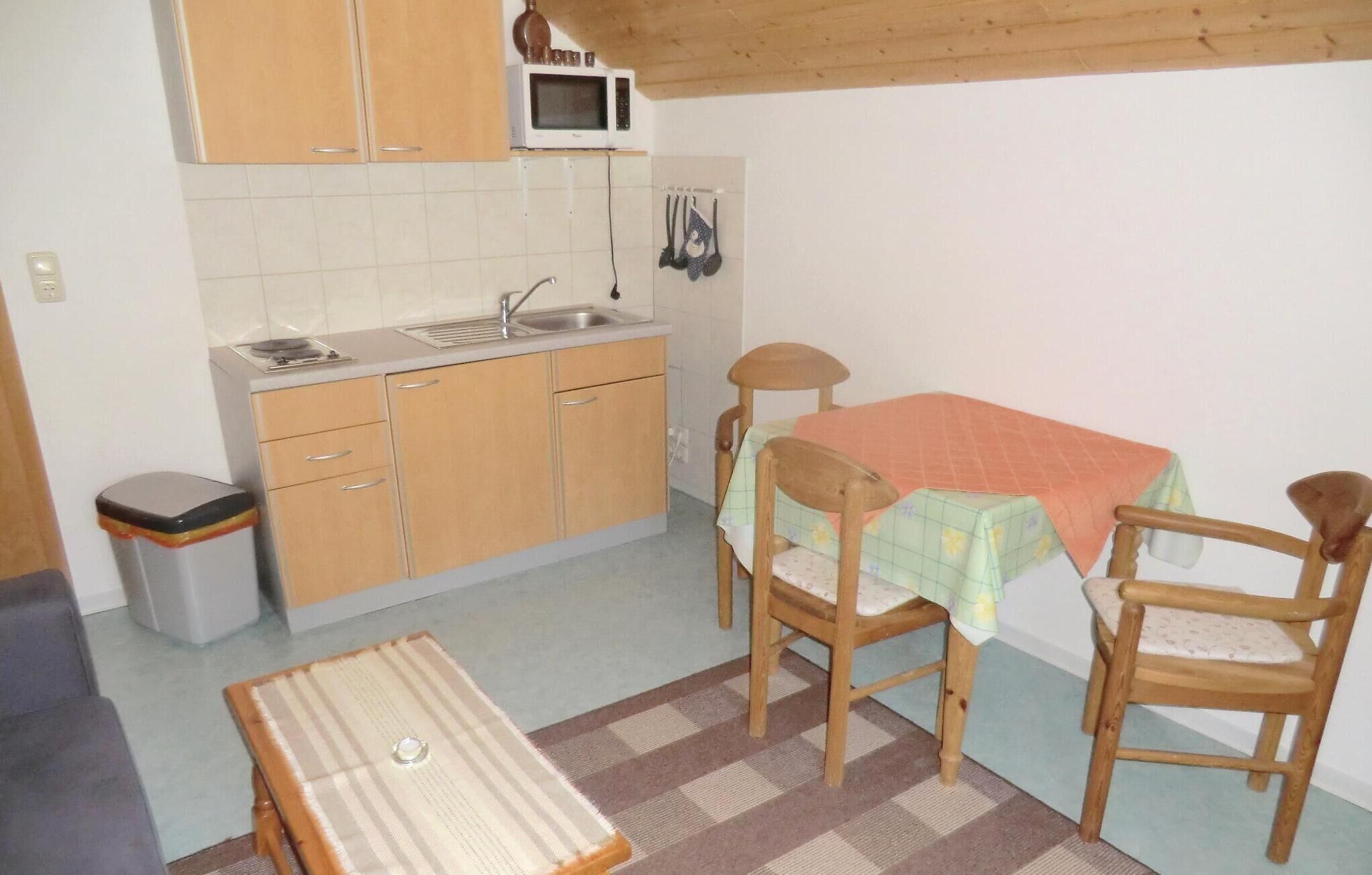 Une cuisine compacte avec des meubles en bois, un four à micro-ondes et un évier. On y trouve une petite table recouverte d’une nappe colorée, ainsi que deux chaises.