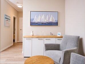 Une pièce comprenant un meuble blanc, un fauteuil recouvert d’un tissu gris, une table basse en bois et un tableau encadré représentant des voiliers accroché au mur.