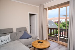 Een moderne woonkamer met een grijze bank, een ronde houten salontafel en een schuifpui naar een balkon.