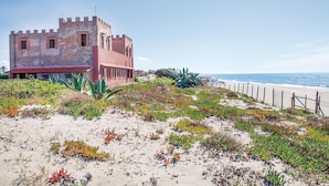 En kystbygning med rødt tag og et tårn, omgivet af vegetation ved stranden og en sti, der fører ned til stranden.