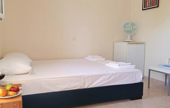 Una cama bien tendida con sábanas blancas, una mesita de noche con una lámpara y un pequeño ventilador sobre un mueble.
