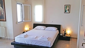 A neatly made bed with white linens and pillows, a bedside table with a wine bottle and glass, and a painting on the wall.