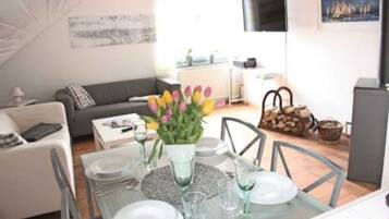 Ein modernes Wohnzimmer mit Essbereich, ausgestattet mit einer Couch, einem Couchtisch und einem Esstisch, der für vier Personen gedeckt ist. Auf dem Tisch stehen Tulpen, und an der Wand ist ein Fernseher angebracht.