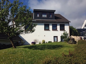 Une maison à deux étages, avec une façade blanche et des tuiles de couleur brune. On y trouve un arbre dans le jardin avant et un petit jardin fleuri.