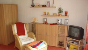 A living room with a wicker chair, a small ottoman, a television on a stand, and wooden shelves with various items. - Vacation apartment Haus am Sonnenhang - Holiday apartment (Bad Sachsa)