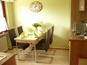 Ein Essbereich mit einem Tisch für zwei Personen, einer Uhr an der Wand und einer Küchenzeile mit Wasserkocher und Gewürzen.