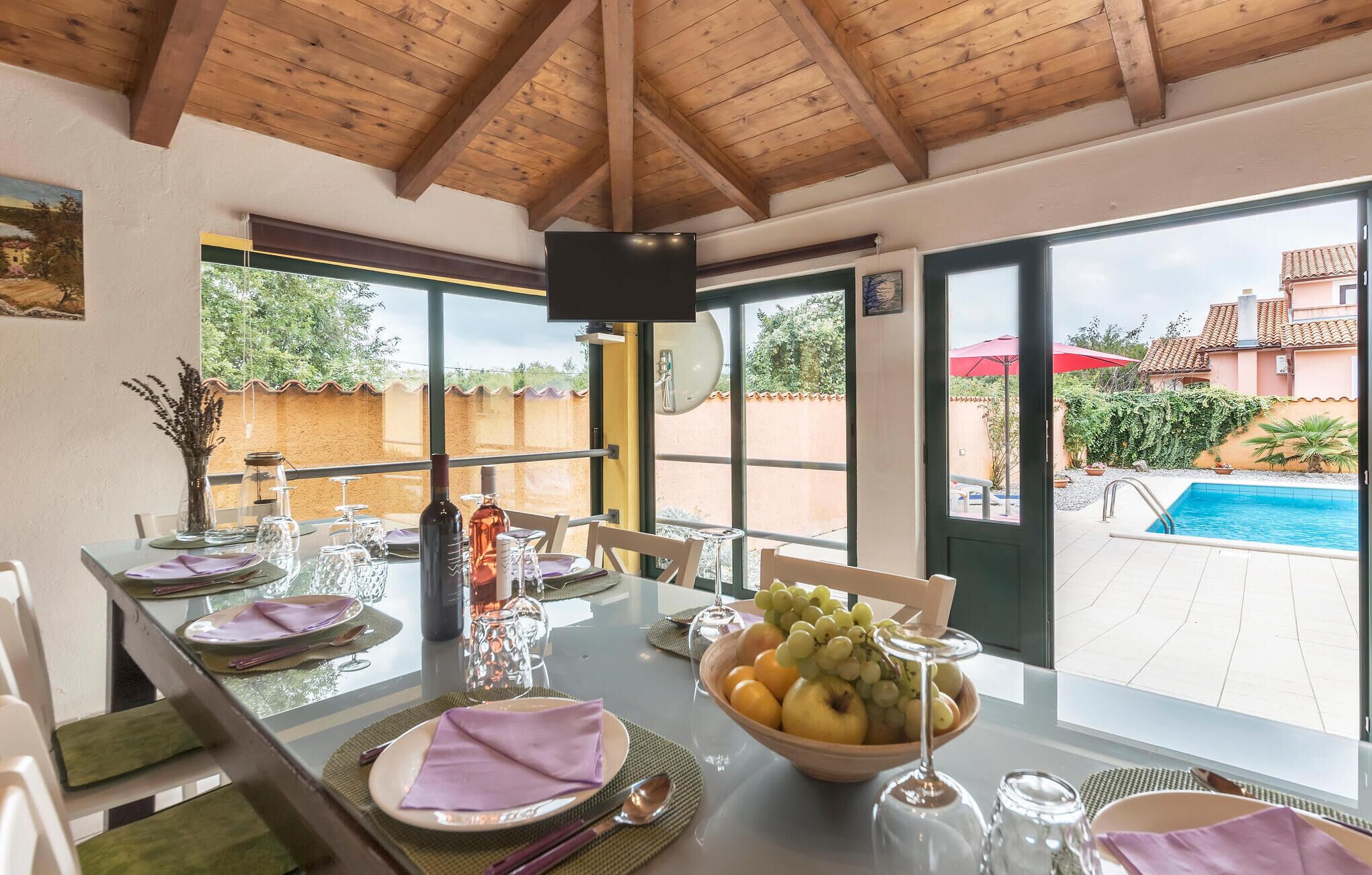Uma área de refeições com uma mesa posta para quatro pessoas, uma cesta de frutas e vista para uma piscina e jardim.