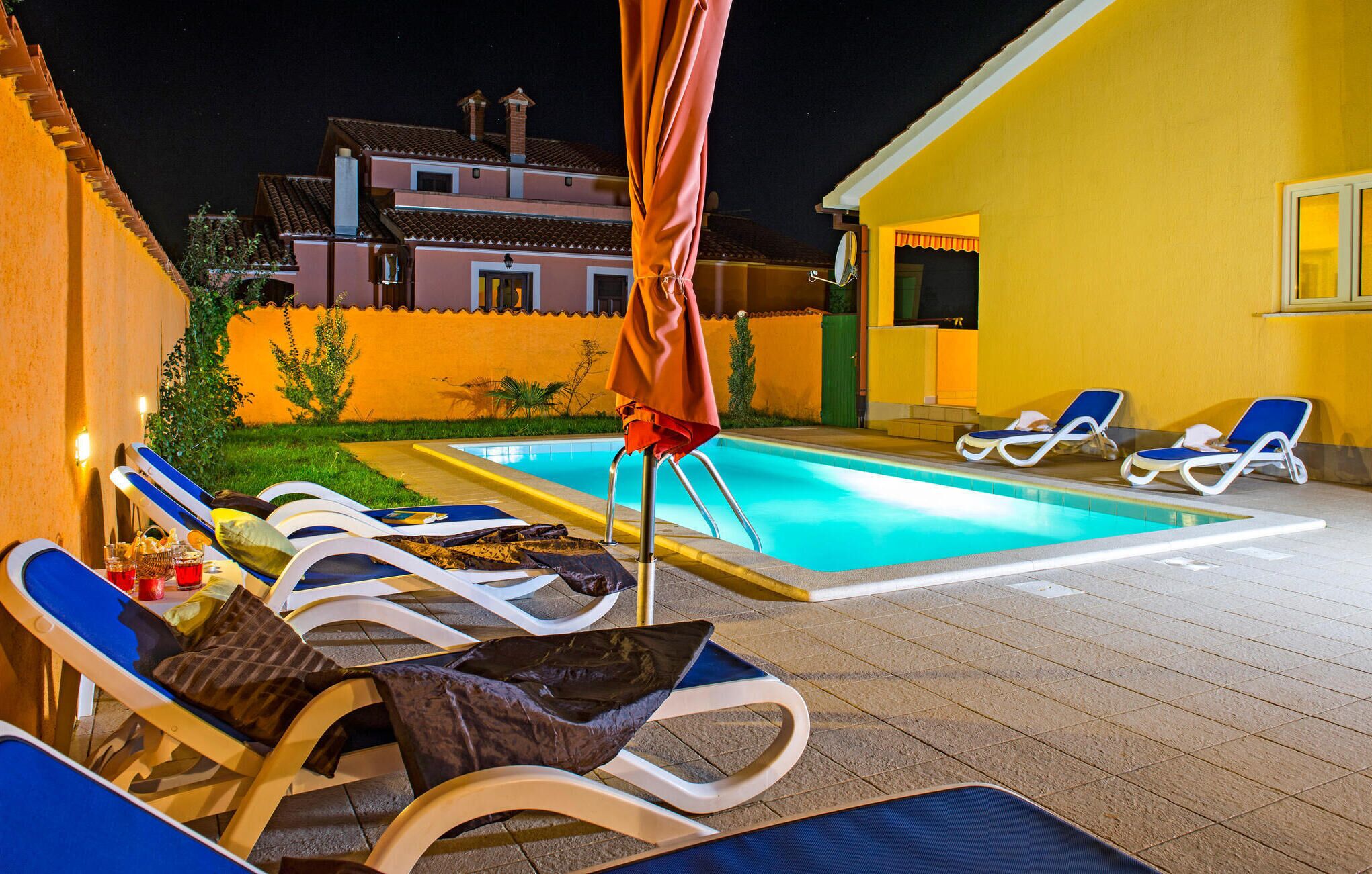 Un área de piscina con sillones de descanso y una sombrilla, un edificio amarillo y una villa al fondo.