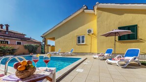 A poolside area with lounge chairs, a table with a fruit basket and drinks, and a yellow building with a window and air conditioning unit. - Amazing home in Labin (Labin)