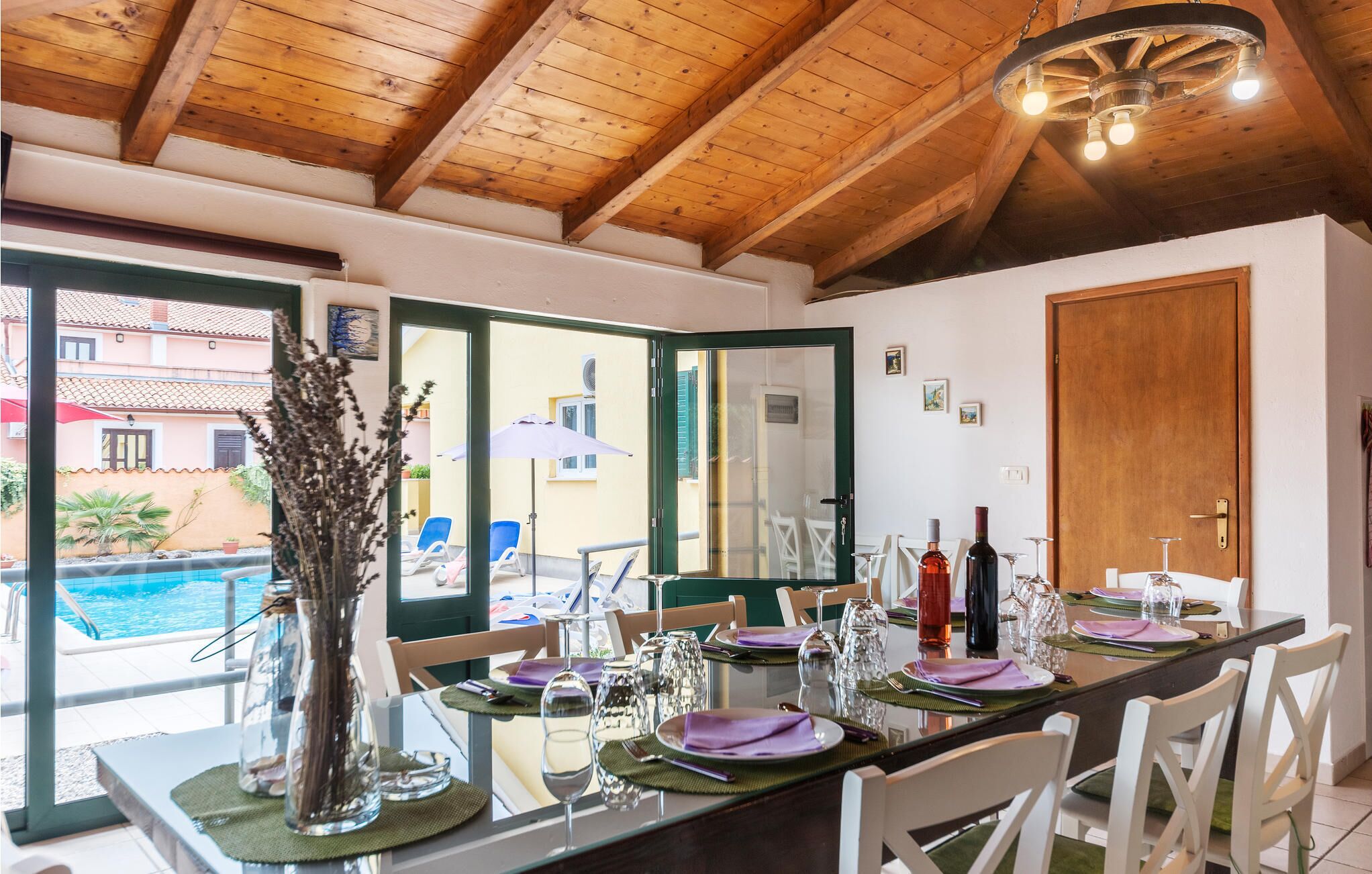 Un comedor con techo de madera, vista a la piscina y una mesa preparada para cuatro personas, con copas de vino y platos.