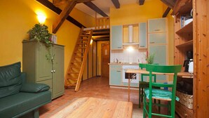 A cozy room with a wooden staircase, a green sofa, a wooden dining table with a chair, and a kitchen area with cabinets and a stove. - Apartment 2 - The Mouse - Vacation at Duckwitz Castle (Behren-Lübchin)