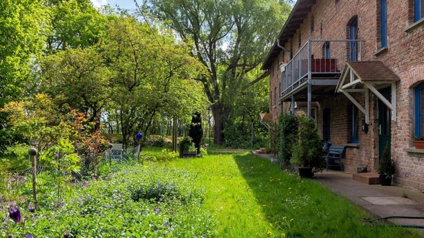 Un bâtiment en briques doté d’un balcon, entouré d’un jardin bien entretenu où poussent diverses plantes et fleurs.