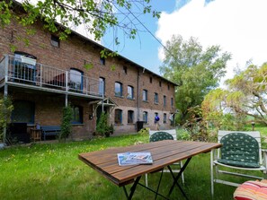 A brick building with a balcony, a wooden table, and green chairs in a garden setting.
