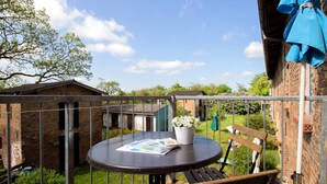 Une table ronde avec un magazine, un banc et un pot de fleurs sur un balcon donnant sur un bâtiment en briques et sur de la verdure.